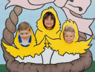 Three children smile in a picture board where they heads pop through the board that's painted like baby chicks Three children smile in a picture board where they heads pop through the board that's painted like baby chicks